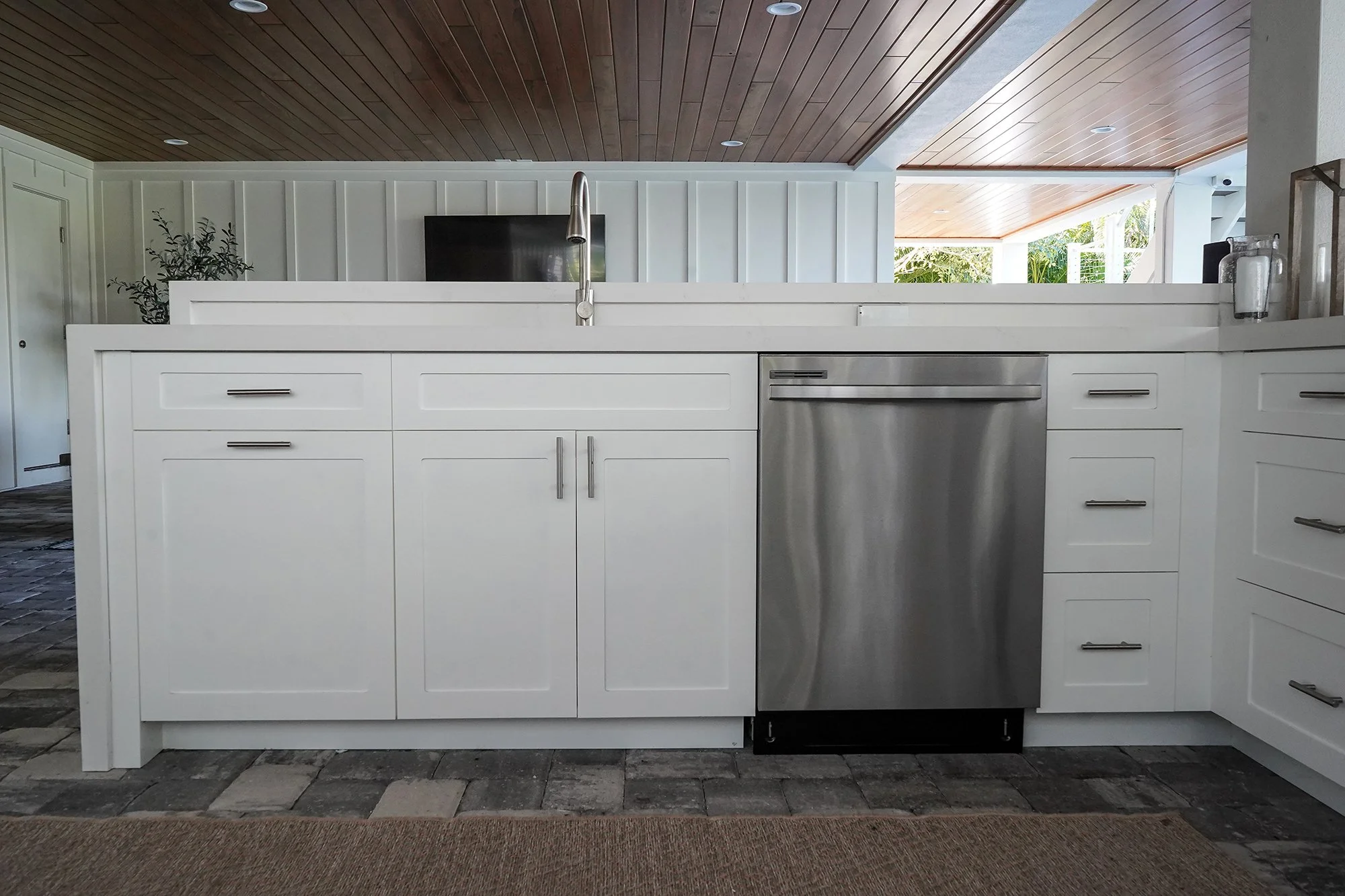 White outdoor kitchen island with a dishwasher under a wood plank ceiling.